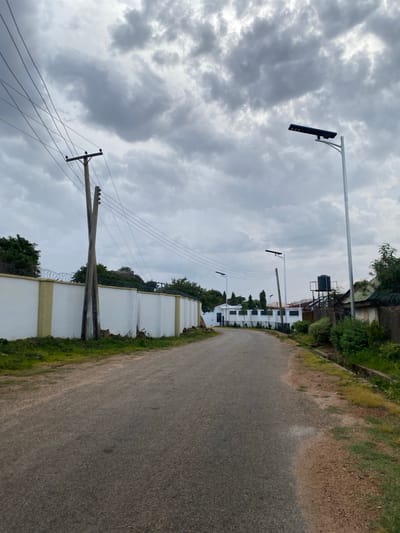 A paved road curves gently to the right in an urban environment under a cloudy sky. On the left side of the road, a white wall topped with barbed wire runs along the property line. A wooden utility pole with multiple wires extends from behind the wall. Further down the road on the left, several modern white buildings with dark-trimmed windows are visible, one with a black-roofed structure. On the right side of the road, a tall, slender metal pole with a solar-powered street light is positioned. Beyond it, another similar street light is seen further down the road. A dark water tank on a metal stand is visible behind a fence on the right, adjacent to a building with a green roof. Vegetation, including trees and bushes, lines the sides of the road and the properties. The overall impression is of a well-maintained residential or commercial area in Barikin Jirga Summa, Nigeria.