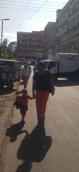 A woman and child walk on a city street with buildings visible. The woman wears a dark top and red pants, holding the child's hand. Cars and a vendor cart are parked nearby. Shadows stretch across the road. The photograph appears rotated, with the horizon tilting upward towards the left.