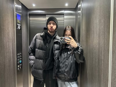 An elevator selfie shows two people. A man on the left wears a black beanie, a black puffy jacket, and a black hoodie with a white logo. A woman on the right wears a black bomber jacket and holds a cell phone with a zebra-print case up to take the photo. The elevator walls are paneled in brown faux wood. The elevator doors are stainless steel, with a button panel visible on the left.