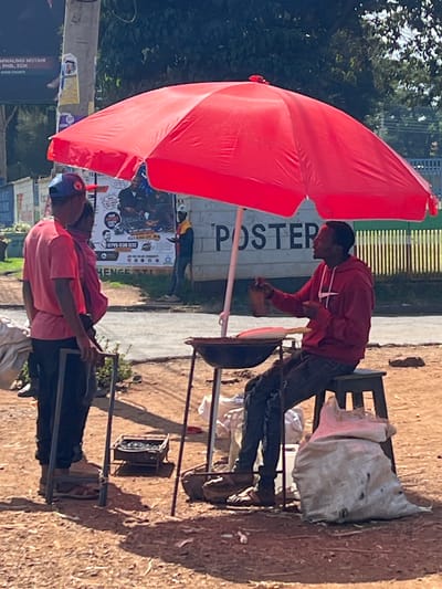 Two people stand by a roadside stand in Nyeri, Kenya. The man on the right is wearing a red hooded jacket and jeans. He is leaning over a cooking surface. The man on the left is wearing a pink shirt, black shorts, and a baseball cap. He is sitting on a stool. A red umbrella provides shade. White bags are placed on chairs behind the men. The background shows posters.
