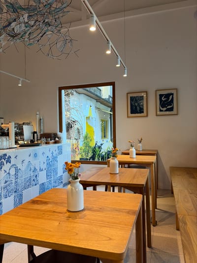 This image shows the interior of a cafe with a row of wooden tables and chairs. The cafe is decorated with blue and white tiles on the walls, and there are two framed pictures on the wall. A window looks out onto a street scene. The overall impression is one of a clean and modern cafe. The cafe is likely located in Ubud, Indonesia, based on the style of decoration and the commonality of cafes in the region.