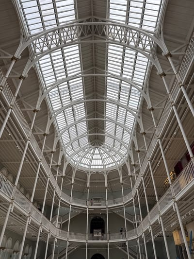 The image shows the interior of the National Museum of Scotland in Edinburgh, United Kingdom, specifically the Grand Gallery. The architecture is characterized by a vast, multi-level atrium with a distinctive glass and iron roof structure that floods the space with natural light. White painted cast iron columns support multiple curved balconies and walkways that encircle the central space, creating a sense of depth and grandeur. Stairs connect the different levels. There are a few individuals visible on the balconies, appearing to be museum visitors. The overall impression is one of an expansive, well-lit, and architecturally significant interior space.