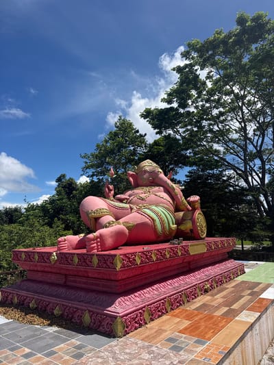 A large, pink, and gold statue of Ganesh reclines on a red, ornate pedestal. The statue is positioned outdoors, with lush green trees and a bright blue sky with white clouds in the background. The pedestal rests on a tiled platform with a mosaic pattern. The overall scene appears to be in a tropical environment. The image was taken in Ko Samui, Thailand.