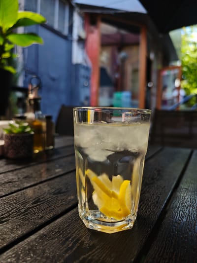 A close-up, eye-level shot showcases a tall glass of water with ice and lemon wedges, placed on a dark wooden table. The glass is positioned slightly right of center, with the water reflecting light and the yellow lemon peel adding a vibrant contrast. In the background, a blur of an outdoor seating area is visible, with hints of greenery, a building with blue siding, and possibly a restaurant or cafe. The lighting suggests it's daytime, possibly late afternoon, with sunlight creating highlights on the table and glass. No people are visible in the frame. The setting appears to be an outdoor cafe or patio in Shumen, Bulgaria.