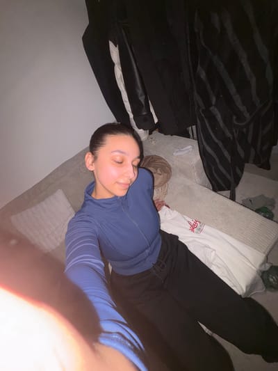 A young woman with her eyes closed is lying on a sectional couch, possibly in a residence in Mönchengladbach, Germany. She is wearing a blue zippered athletic top and black sweatpants. The couch is covered in a textured fabric and has a white decorative pillow. Behind her, there are several dark-colored garments hanging on what appears to be a coat rack or similar structure. A white t-shirt with red lettering and a brown duffel bag are also visible on the couch. The lighting suggests an indoor setting, likely during the evening or night.