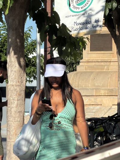 A woman wearing a white visor and a mint green crochet dress is looking down at her phone. She is holding a white plastic bag in her left arm and has several gold bracelets on her right arm. Behind her, a banner reads "NIGHT MARKET SINCE 1975" and "Mercadillo del Puerto de Ibiza." Bicycles are parked to the right of the woman, and a tree trunk is to her left. The setting appears to be a street market in Eivissa, Spain.