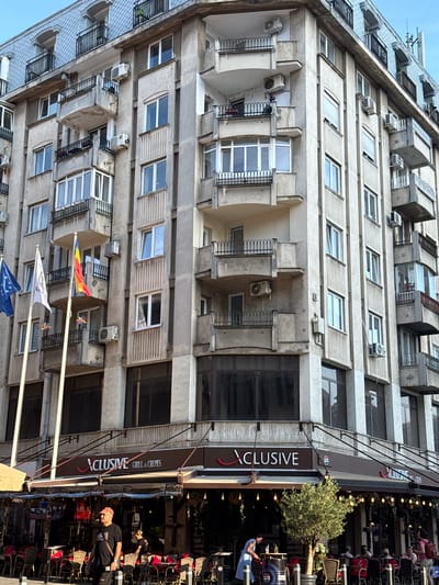The image shows a street view in Bucharest, Romania, focusing on the exterior of a building with a restaurant named "XCLUSIVE GRILL & CREPES" on the ground floor. The building is a multi-story residential and commercial structure with a facade of worn concrete and balconies on multiple levels. Many of the balconies have air conditioning units attached. Flags are visible on poles in front of the building, including the Romanian flag and the European Union flag. The restaurant has outdoor seating with tables and chairs, some covered by awnings. People are visible at tables and walking past the restaurant. A man in a red cap and black t-shirt is walking in the foreground, and a woman in a blue dress is working near a table inside the restaurant. The overall scene depicts a typical urban environment with a mix of residential and commercial activity.