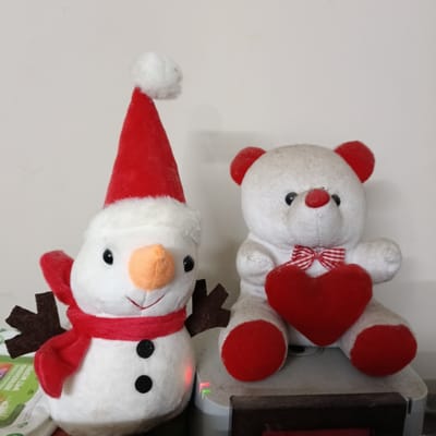 A photograph taken indoors in Bengaluru, India shows two plush toys: a snowman and a bear. The snowman, on the left, has a white body, a red Santa hat with a white pom-pom, a red scarf, a carrot nose, black button eyes and mouth, and brown felt antlers. The bear, on the right, is white with red ears, a red nose, red feet, and a red and white striped bow tie. It is holding a red heart. Both toys are placed on a surface, which is slightly dirty. A package of wet wipes is partially visible on the left side of the frame. The background is a plain off-white wall.