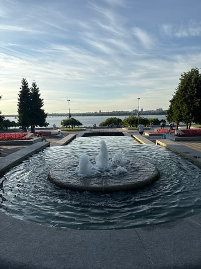 The image depicts a waterfront promenade in Dnipro, Ukraine, bathed in the soft light of what appears to be late afternoon or early evening. A prominent feature is a circular fountain with water jetting upwards, creating ripples that cascade into the surrounding basin. The water's surface is textured with numerous small bubbles and waves, reflecting the sky.
To the left of the fountain, a paved walkway runs alongside a body of water, likely the Dnipro River, which stretches across the background. A distant cityscape is visible on the opposite bank, with buildings and a bridge suggesting an urban environment.
The promenade is lined with trees and flowerbeds filled with red flowers, adding color to the scene. Several people are scattered along the walkway, some seated on benches or near the flowerbeds, while others stroll leisurely. Their presence suggests a relaxed atmosphere, with individuals enjoying the waterfront ambiance.
The sky above is a canvas of wispy white clouds against a pale blue backdrop, indicating fair weather. A few lampposts are visible along the promenade, hinting at the infrastructure supporting this public space. The overall mood is serene and tranquil, capturing a moment of calm enjoyment in an urban riverside setting. No visible text is present in the image.