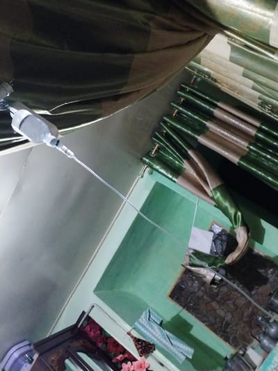 The image displays an indoor scene, likely a room within a residential or medical care environment. A white-covered intravenous (IV) fluid bag or bottle is suspended in the upper left, with a transparent plastic tube extending diagonally downward and to the right. Above this, a large section of draped, olive-green and beige striped fabric, possibly a curtain, is visible. To the right, a pleated curtain of identical green and beige striped fabric is affixed to a horizontal fixture. The walls exhibit a light green painted finish. Towards the bottom right, a segment of dark, textured patterned flooring or a mat is observed, bearing a small white rectangular object. In the lower left, a dark wooden cabinet featuring gold detailing and glass panels holds pink and red artificial flowers. Adjacent to the cabinet, a folded blue and white patterned cloth, several small metallic pots or containers, and a decorative object resembling a butterfly are present on a surface. No individuals are depicted. The visual information within the image does not contain identifiable geographical markers to confirm its location as Limbur, Uzbekistan.