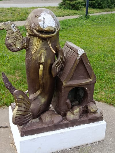 This image shows a weathered, bronze-colored sculpture of a mythical creature resembling a seal with a fish tail, accompanied by a small dog house. The sculpture is located in the city of Elovo, Russia, situated on a concrete base with a white plaster coating. The creature has a large, round head with distinct eyes and a wide mouth, and it is positioned next to a brown dog house that has an arched opening. The sculpture appears to be outdoors, with a grassy area and a paved walkway visible in the background. The overall condition of the sculpture suggests it has been exposed to the elements, with some areas of peeling paint and wear.