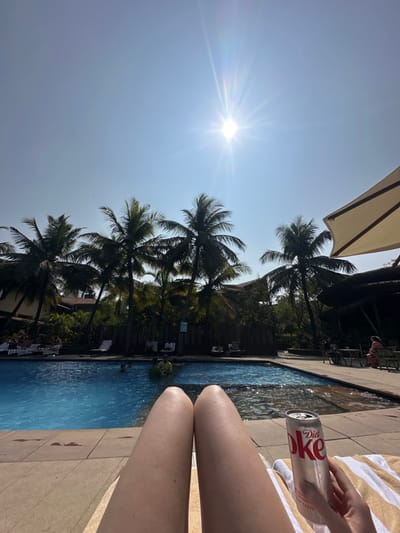 A sun-drenched scene features a person relaxing poolside. Palm trees line the horizon against a bright sky with the sun shining prominently. The clear blue pool reflects the surroundings. A Diet Coke can is held in hand, and legs extend toward the water's edge. A partially visible sunshade and a striped towel complete the relaxing summer setting.