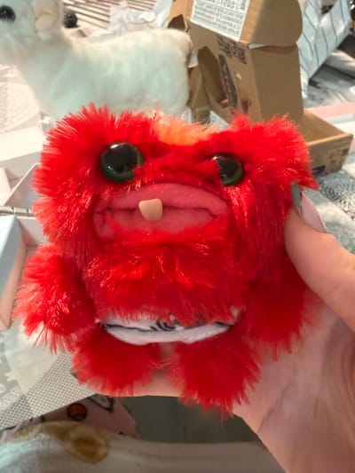 A person's right hand, with light-colored nail polish, holds a small, red, fluffy stuffed toy in the foreground. The toy has large, glossy black eyes, an open mouth revealing a pink interior, and a single off-white protruding tooth. A segment of black and white patterned fabric is visible on the toy's underside. The background is blurred, showing a white, fluffy, animal-shaped toy to the left and an open cardboard box with Asian script to the right. The scene takes place indoors, within a domestic setting in Raanana, Israel.