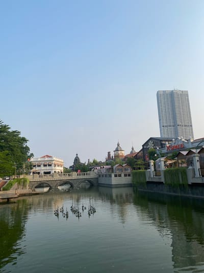 The image shows a waterfront scene with buildings and a tall skyscraper reflected in the water. The buildings have a European architectural style, with domed roofs and arched windows. Some of the buildings are adorned with hanging plants. A large, modern skyscraper stands in the background, its reflection distorted in the rippling water. The sky is a clear, pale blue, suggesting it is daytime, likely morning or late afternoon due to the soft light. The water appears calm, with gentle ripples.
The scene is set in a developed urban area, with the presence of both historical-style architecture and a contemporary skyscraper indicating a mixed-use environment. There are no people visible in the image. The overall mood is tranquil and picturesque, emphasizing the interplay between the built environment and its reflection.
Visible text includes "XGRAND" in orange lettering on a sign above one of the buildings, and another partially visible word "CORPO" on a building facade across the water. The location context is East Jakarta, Indonesia.
