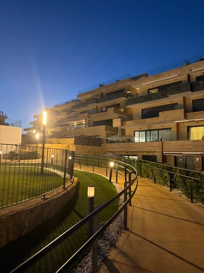 The image depicts a modern multi-story building complex at dusk in Finestrat, Spain. The building features a terraced design with light-colored stone facades and dark-tinted windows. Balconies with glass railings are visible on various levels, and some appear furnished with outdoor seating. A curved pathway with metal handrails leads through the complex, bordered by artificial turf and decorative stone. Several modern, rectangular outdoor lights illuminate the pathway and surrounding areas. In the background, the deep blue sky transitions to a lighter shade near the horizon. A single individual is visible walking on the left side of the image, near a white building structure. The overall scene conveys a sense of contemporary urban living with an emphasis on outdoor amenities and architectural design.