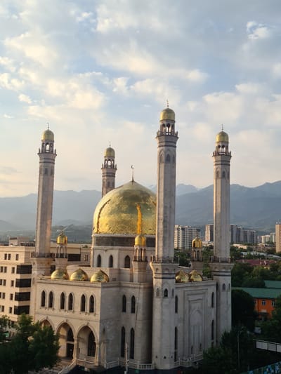 The image depicts the Central State Mosque in Almaty, Kazakhstan, bathed in the warm glow of what appears to be either sunrise or sunset. The mosque, a magnificent architectural edifice, is characterized by its dominant golden dome, reflecting the sunlight. Surrounding the central dome are four towering minarets, also topped with smaller golden domes, accentuating the mosque's grandeur. The facade of the mosque is constructed from light-colored stone, featuring arched windows and intricate detailing.

In the background, a backdrop of majestic mountains, possibly part of the Tien Shan range, rises against a sky filled with scattered clouds. The city of Almaty is visible, with a mix of modern high-rise buildings and lower-rise structures interspersed with lush greenery. The overall atmosphere conveyed is one of peace and serenity, with the mosque standing as a prominent spiritual landmark within the urban and natural landscape. There are no people visible in the image, and the focus is solely on the architectural and environmental aspects of the scene.