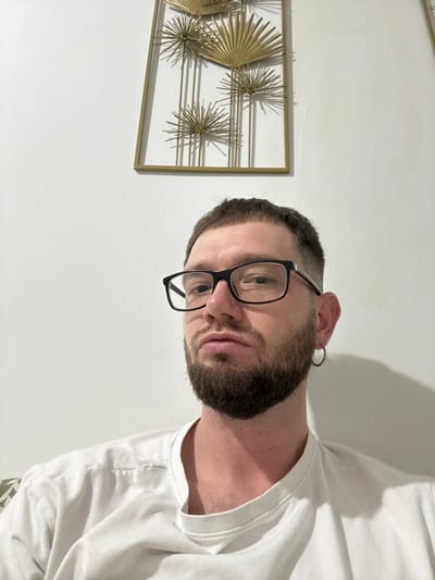 An adult male is depicted from the chest up, centered in the lower portion of the frame, against a plain white wall. He has short, dark hair, a trimmed beard, and is wearing black-framed rectangular glasses. A single silver-colored hoop earring is visible in his left ear. He wears a plain white crew-neck t-shirt. His gaze is directed forward towards the camera, with a neutral facial expression. In the upper right quadrant of the background, a rectangular piece of gold-framed wall art is mounted on the white wall. The art features stylized golden elements resembling fan-like leaves and spiky starbursts, interconnected by vertical lines. The wall exhibits slight imperfections and texture. The specific location within Madrid, Spain, cannot be determined from the visual information present in the image.