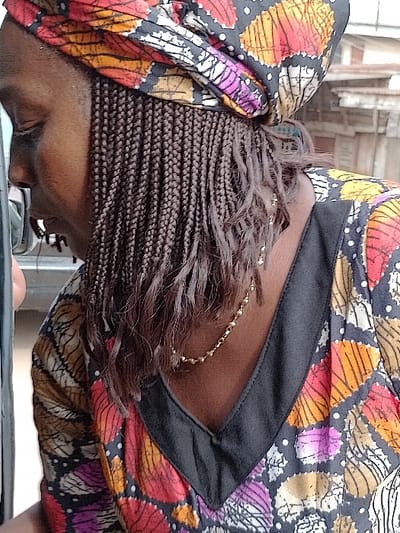 A person with dark skin and braided hair adorned with a colorful headscarf bends forward. The headscarf features a mix of orange, red, pink, and white patterns. The person wears a blouse with a similar colorful pattern and a black collar. A delicate gold necklace is visible at the base of the neck, peeking out from beneath the hair. The focus is on the back of the head and neck, partially obscured by the braided hair.