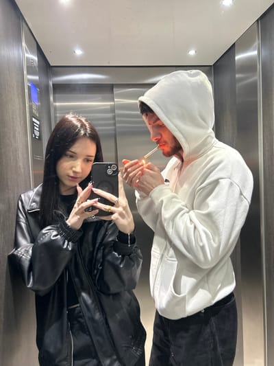 Within an elevator in Cachan, France, two individuals are captured in a mirror selfie. The female subject, positioned on the left, has long dark hair and wears a black leather-style jacket over a dark top. She holds a dark-colored smartphone with a triple camera array in her right hand, pointed towards the mirror, seemingly capturing the image. Several rings are visible on her fingers, and a silver-colored bracelet is on her left wrist. Her gaze is directed towards the phone screen. The male subject, positioned on the right, wears a white hooded sweatshirt with the hood up and dark trousers. He holds a lit cigarette in his mouth with his left hand and a lighter in his right hand, actively igniting the cigarette, with a bright orange flame visible. The light from the flame casts an orange glow on his face. The elevator's interior features reflective, brushed metal wall panels and recessed ceiling lights. A control panel with illuminated buttons is visible on the left wall.
