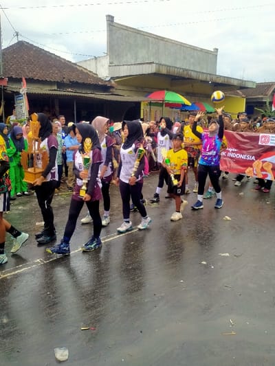 A daytime image depicts a procession on a wet asphalt road in Mojogedang, Indonesia, under an overcast sky. The foreground and midground are occupied by multiple individuals, predominantly young females, dressed in volleyball jerseys and athletic wear, many wearing hijabs. Several participants carry trophies of varying sizes; specifically, one young female in a white and maroon jersey (number 18) carries a gold trophy, and another female carries a large wooden trophy. A female in a vibrant purple, pink, and blue jersey (number 1) is captured mid-action, setting a yellow and blue volleyball overhead. A young boy in a yellow jersey (number 17) holding a gold trophy is also visible. The wet road surface reflects the overcast sky and the individuals. In the background, a crowd of spectators or additional participants observe the procession. Buildings line the street, including a traditional structure with a tiled roof on the left and a larger, more modern concrete building behind it. A colorful, multi-paneled parasol is visible in the background, along with a partially obscured red banner displaying white text, including "PERSIK INDONESIA" and "80 tahun." Overhead wires are visible against the sky. Small debris, including a white object and a thin stick, is present on the road surface.