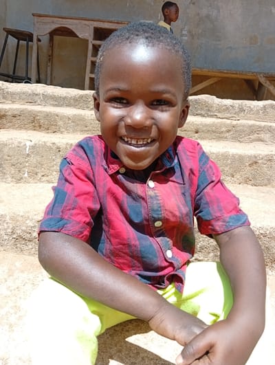 An outdoor scene in Rido, Nigeria, features a young child with dark skin and short, dark hair, seated on rough concrete steps under direct sunlight. The child wears a red, black, and dark-colored plaid short-sleeved collared shirt with white buttons, and bright yellow-green trousers. He is smiling directly at the camera, displaying teeth, with his hands resting in front of him. Behind the foreground child, a series of light-colored concrete steps ascend. In the midground, a dark-colored metal three-legged stool and a light-colored wooden desk with a curved front and internal shelving are positioned against a light-colored, textured wall. A second, smaller child with dark skin and a light-colored shirt is partially visible in the upper right background, standing further back and looking to the right, away from the camera.