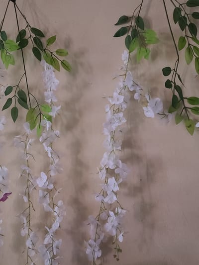 The image shows artificial floral and foliage garlands against a plain, light brown wall. The garlands are arranged horizontally in two rows. The top row features a garland with small white flowers interspersed with green leaves. The bottom row displays a similar arrangement. The background is a simple wall with a slightly textured surface and a neutral color. There are no people, subjects, objects, actions, or interactions depicted in the image beyond the garlands and the wall. There is no specific evidence suggesting the image's location is Nyeri, Kenya.