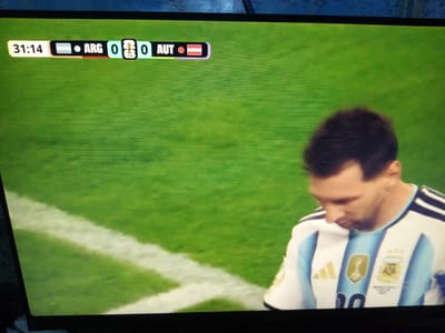 A screen displays a football match score. The time is 31:14. Argentina (ARG) has 0 goals, and Austria (AUT) has 0 goals. The match is being played on a green grass field with white lines. A person wearing an Argentinian football jersey with the number 10 is visible in the lower right portion of the screen. The screen is positioned within a room in Chirakchi, Uzbekistan.