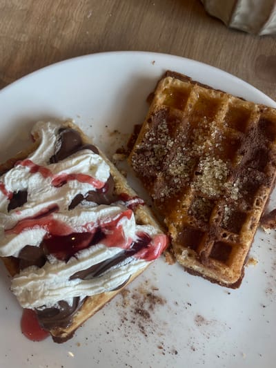 Two waffles are presented on a white plate. The waffle on the left is topped with whipped cream, chocolate spread, and a red syrup. The waffle on the right is topped with a syrup, cocoa powder, and coarse sugar crystals. The plate is resting on a wooden surface. The image is a close-up shot, with the focus on the food. The scene is indoors, likely a cafe or restaurant. No people are visible in the image. The location is not identifiable as Budapest, Hungary from the visual information provided.