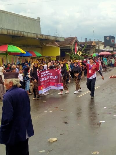 A forensic examination of the image reveals a street scene in Mojogedang, Indonesia, depicting a procession or parade under an overcast sky. The asphalt road surface is wet, showing reflections and scattered litter, including paper, plastic, and organic debris.
In the immediate foreground, facing away from the viewer and slightly to the left, is an elderly man with grey hair and a beard, wearing a dark blue long-sleeved garment and a dark peci (cap). He observes the ongoing activities.
Midground features a group of approximately four individuals, predominantly women, carrying a large red banner horizontally across the street. The banner bears white text, prominently displaying "KARNAVAL KEMERDEKAAN XALITELU" and smaller text below, including "2023" and motifs resembling the Indonesian flag. The women carrying the banner wear black tops, patterned batik-style bottoms, and gold-colored headpieces. To the right of the banner, a man in a red and white short-sleeved shirt with an apparent Indonesian flag emblem and a lanyard with an ID card walks with his arms slightly outstretched, looking upwards and to his right.
The background is densely populated with spectators lining both sides of the street. On the left, multiple colorful market stalls with red, green, and blue umbrellas are visible, under which crowds gather. Beyond these, a mix of single and two-story buildings, characteristic of commercial or residential structures, line the street. A light-colored, multi-story building with a bare concrete upper floor is prominent on the far left. On the right, further buildings with brown tiled roofs are visible, along with a warning sign (yellow diamond with an exclamation mark) affixed to a pole amidst foliage.