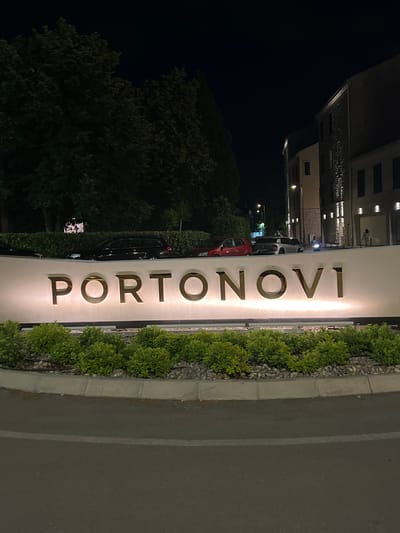 This is a nighttime image taken in Kumbor, Montenegro. The primary subject is a large, illuminated sign that reads "PORTONOVI" in gold, three-dimensional letters against a white background. The sign is mounted vertically and is bordered by neatly trimmed green bushes on the right and a stone curb with gravel on the left.
In the background, the scene is dark, suggesting it is night. To the left of the sign, there is a glimpse of a modern building with illuminated windows, indicative of a resort or hotel. Further left, there are trees and foliage, also dark against the night sky. A few cars are visible parked behind the sign and bushes, illuminated by the sign's light. One car is red, another is gray, and a third is black, with its outline clearly visible. The ground appears to be an asphalt road with a white line marking.
There are no people clearly visible or engaged in any specific activity. The overall impression is one of a well-lit entrance to a resort or development at night, emphasizing its name and upscale appearance. The lighting on the sign and building suggests a warm and inviting atmosphere, despite the darkness of the surrounding environment. The weather appears calm.