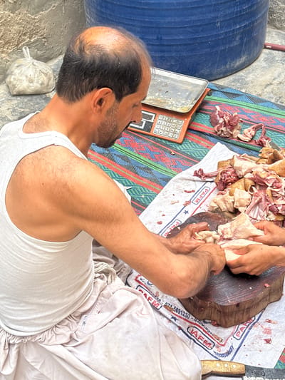 A man in a white tank top sits on a white cloth on a woven mat, cutting meat with his hands on a wooden block. To the left is a blue plastic barrel and an orange scale. A plastic bag sits on the ground next to the barrel. The scene is in Quetta City Tehsil, Pakistan.