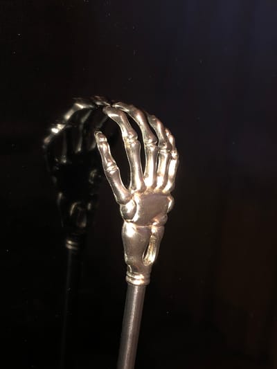 A metallic skeleton hand is reflected in a dark, possibly glass, surface. The hand is positioned on a slender, metallic rod, suggesting it might be part of a decorative item, utensil, or prop. The background is entirely black, providing a stark contrast to the metallic sheen of the object. The reflection is clear and mirrors the object precisely, indicating a smooth, polished surface. The lighting emphasizes the reflective qualities of the metal. There are no discernible features of Baku City, Azerbaijan in the image, nor any people, actions, or interactions.