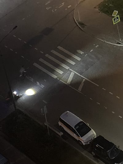The image shows an aerial view of a street at night in Misaylovo, Russia. A pedestrian crossing is visible in the center of the frame, marked by white stripes on the asphalt. To the left of the pedestrian crossing, a single vehicle with its headlights on is positioned on the road. Further down the road, two cars are parked, one white and one black. A pedestrian crossing sign is visible on the right side of the image, near a grassy area. The scene appears to be a typical urban street environment at night, with streetlights providing illumination.