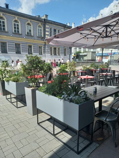 The image shows an outdoor seating area of a cafe in Yekaterinburg, Russia, likely on a sunny day. In the foreground, there are several large, modern grey planters filled with a variety of greenery. These planters are supported by black metal frames, and they are positioned on a light grey brick-paved sidewalk. Behind the planters, tables and chairs for the cafe are visible. Some of the tables are round and red, while others are rectangular and dark grey. The chairs are dark metal with a distressed finish. A large, light-colored umbrella, adorned with lights, covers a portion of the seating area. In the background, a multi-story building with a light grey facade and arched windows is visible. People are walking in the background, suggesting the area is public. The overall impression is a well-maintained outdoor cafe space in an urban environment.