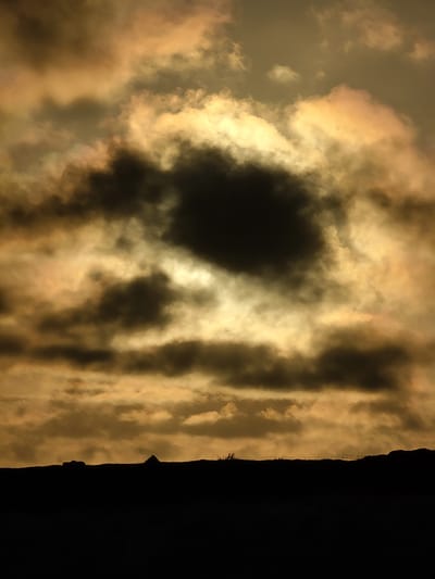 The image depicts a dramatic sky over Quarteira, Portugal, during what appears to be sunset or sunrise. The sky is filled with a mix of dark, ominous clouds and brighter, golden-hued clouds that are illuminated by the low sun. The light filtering through the clouds creates an ethereal and somewhat turbulent atmosphere.
On the right side of the image, a dark silhouette rises, suggesting a landmass, possibly cliffs or a man-made structure. The jagged edge of this silhouette contrasts sharply with the softer, more diffuse forms of the clouds. Tiny, almost indiscernible shapes near the top of the silhouette might be trees or man-made structures like pylons.
The overall impression is one of natural beauty with an element of mystery or foreboding due to the intense contrast between light and dark. The colors are rich, with warm oranges and yellows blending into deep grays and blacks. There is no visible text in the image. The scene evokes a sense of awe at the power and spectacle of nature.