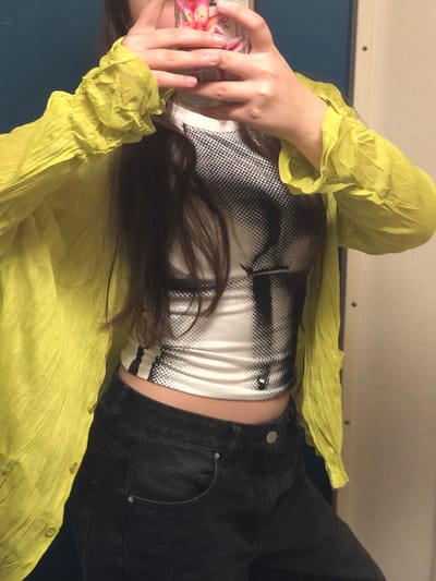 A person lies on their side, facing away from the camera. The individual wears a cropped white top with a black and white graphic print, black jeans, and a sheer, bright yellow button-up shirt. The person is holding a cell phone up to their face. The background appears to be a train car interior with a dark blue wall. The image is slightly tilted.