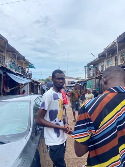 In Ogidi, Nigeria, two young men are captured in a medium shot on a street, with a gray car in the foreground on the left. The man on the left is facing the camera, wearing a white t-shirt with a colorful bear graphic and black and white patterned pants. He has a red cloth draped around his neck. The man on the right is seen from the back, wearing a brightly striped shirt. They appear to be interacting, with both men holding their hands in front of them, possibly engaged in a conversation or a handshake. The background reveals a bustling market scene with stalls lining both sides of the street, displaying goods under awnings. Several other individuals are visible in the background, some browsing or walking through the market. The sky is overcast, suggesting a cloudy day. The overall environment depicts a lively urban market atmosphere in Ogidi.