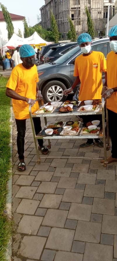 In Giginyu, Nigeria, three individuals are positioned near a dark gray SUV. Two of the individuals, wearing blue hair coverings and orange t-shirts, are actively engaged with a metal serving cart laden with meals. The cart, positioned in the foreground, displays multiple platters of food, each containing rice, meats, and various accompaniments. Both individuals are wearing clear plastic gloves, indicating they are handling the food. The third individual, partially visible on the left, also wears a blue hair covering and an orange t-shirt, and appears to be observing the scene. The environment includes a paved walkway, green grass, and a building under construction in the background, with a white tent structure visible to the left.