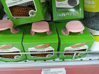 A retail display shelf in a store in Angren, Uzbekistan, holds green cardboard boxes of dish scrubbers. Each box features the brand name "Garnet" and text in Cyrillic. The front of the boxes has a cutout through which the scrubbers, made of metallic gold-colored wire, are visible. Some of the boxes also have pink plastic handles protruding from the top. The shelf is made of metal wire. In the foreground, a price tag with the "fresh" supermarket logo and Cyrillic text is partially visible.