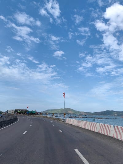 The image depicts a coastal road in Hòa Hiệp, Vietnam, under a bright blue sky with scattered white clouds. The asphalt road curves to the left, marked with white dashed lane lines. To the left of the road, a dark grey barrier runs along the edge. To the right, a concrete barrier with alternating red and white diagonal stripes separates the road from a body of water, likely a bay or inlet. This water area contains numerous aquaculture structures, possibly fish or oyster farms, and several small boats are visible among them.
In the mid-ground, a tall pole with a prominent red flag flies on the right, near the water's edge. Further down the road on the left, a cluster of buildings, some appearing commercial, are visible. A dark-colored vehicle is partially visible on the far left, parked or in motion. Along the right side of the road, yellow directional signs are present. Across the water, green, undulating hills or mountains form the distant horizon. No individuals are clearly discernible in the scene.