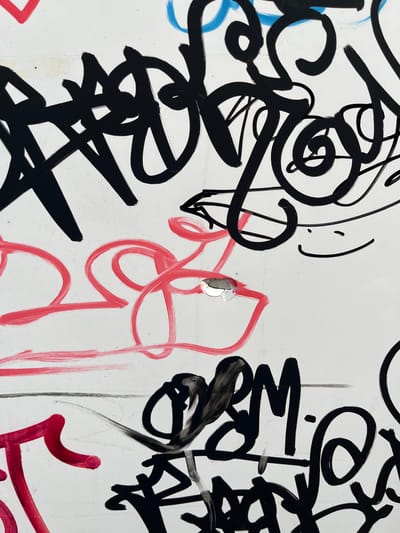 The image shows a close-up of graffiti on a white surface, likely a wall or a metal panel. The graffiti consists of black, red, and blue markings in a stylized, calligraphic manner, typical of urban art. There is a small, circular hole with a reflective surface in the center of the image, possibly a rivet or damage to the surface. The overall impression is of a vibrant, albeit slightly chaotic, display of street art. The location information provided, Podostrog, Montenegro, indicates an urban setting where such graffiti is commonly found.