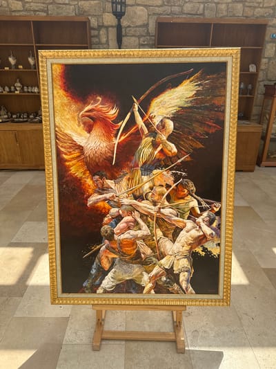 The painting, displayed on a wooden easel, depicts a dramatic scene featuring a mythological phoenix and a group of figures engaged in combat. The phoenix, rendered in vibrant reds and oranges, dominates the upper left portion of the artwork, its wings outstretched and engulfed in flames. Below and to the right of the phoenix, a group of muscular, semi-nude figures, some holding bows and arrows, are shown in various states of action and struggle. The figures are depicted with dynamic poses and intense facial expressions, suggesting a fierce battle. The background of the painting is dark, further emphasizing the illuminated figures and the phoenix. The painting is enclosed in an ornate golden frame.

The painting is located in a room with stone walls and wooden shelving units. On the shelves to the left of the painting are various decorative ceramic vases and bowls. To the right, a wooden cabinet and a display case can be seen. The floor is tiled with large, light-colored stone tiles, with sunlight casting geometric shadows from an unseen source. The overall setting appears to be a museum or gallery. The city of Neofit Rilski, Bulgaria, is not visibly represented in the image.
