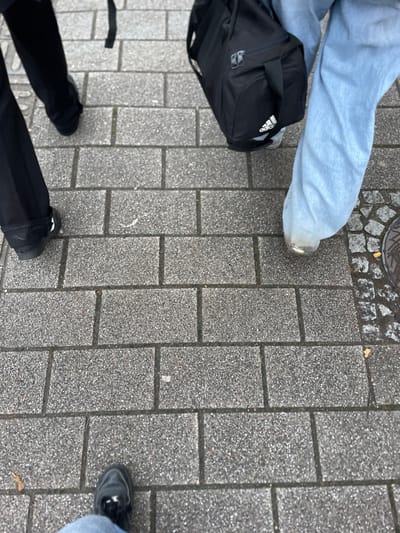 The scene depicts a ground-level view of three individuals' lower extremities standing on a paved surface in Schiltigheim, France. The environment consists primarily of grey, rectangular interlocking pavers forming a sidewalk or pedestrian area. To the far right, a section of smaller, irregularly shaped cobblestones and a circular metal manhole cover are visible.
Three sets of feet and lower legs are present:
1. In the immediate foreground, the lower portion of blue jeans and a black shoe are partially visible, angled left.
2. To the mid-left, a person wears black trousers and black shoes, with one leg visible from mid-calf and the other partially obscured.
3. To the mid-right, a person wears light blue, wide-leg jeans and light-colored shoes, with the bottom hem of the jeans appearing worn or scuffed. This person carries a black Adidas duffel bag with a white logo, positioned to the right of their leg.
Scattered small debris, possibly dried leaves or light specks, are sparsely distributed across the paving. No explicit actions or interactions beyond standing are discernible.