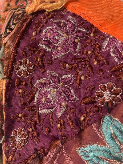 The image displays a close-up view of intricate fabric embroidery, featuring floral motifs and decorative beads. The dominant colors are deep purple, orange, and teal, with accents of silver and brown thread. The fabric appears to be part of a textile artwork or garment, possibly a collage of different materials given the varying textures and colors visible. There are no people, city landmarks, or environmental details that would indicate a location in Sabadell, Spain, or any specific geographical setting. The scene is purely focused on the detailed craftsmanship of the embroidery.
