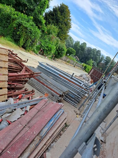 The image depicts a construction site with stacks of scaffolding poles, wooden planks, and construction materials. The scene is outdoors, with greenery and trees in the background under a partly cloudy sky. The ground is covered in dirt and gravel. Specifically, the image captures a pile of red-painted metal scaffolding pipes on the left, adjacent to a stack of wooden pallets. To the right of the red pipes are neatly stacked metal scaffolding planks. In the foreground, there are also red and blue painted wooden planks, possibly used as formwork. A large roll of white material, possibly fabric or tarp, is visible further back in the scene, behind the scaffolding. Further to the right, construction scaffolding poles are visible, and part of a building's wall is in the far right. The location is identified as Settimo Torinese, Italy. There are no people or animals visible in the image, and no specific actions or interactions are depicted. The overall impression is one of an organized, albeit partially completed, construction area.
