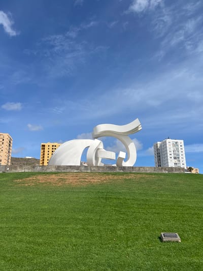 The image shows the "Monumento al Atlante" sculpture in Las Palmas, Spain, set against a blue sky with scattered clouds. The sculpture is a large, white abstract structure. Behind the sculpture are several buildings, including a tall white apartment building and other structures of varying heights. To the right of the sculpture is a green grassy area separated by a stone retaining wall. A plaque or marker is visible within the grassy area.