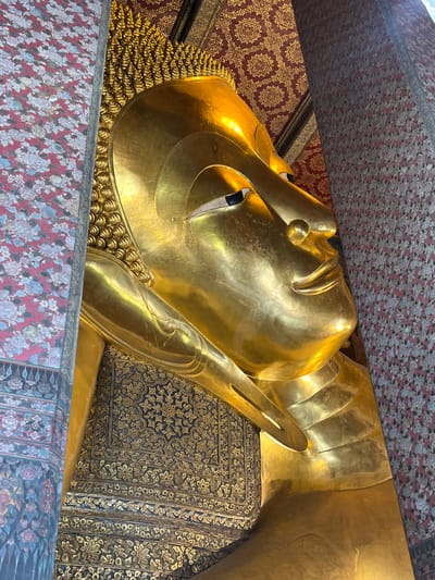 The image shows a close-up of a large, golden, reclining Buddha statue. The statue is highly detailed, with intricate carvings on its hair and body. The background consists of richly decorated walls with floral patterns. This statue is located in Wat Pho, also known as the Temple of the Reclining Buddha, in Bangkok, Thailand. The lighting suggests the image was taken indoors, likely within the temple complex. The focus is on the serene expression of the Buddha's face and the golden sheen of its body.