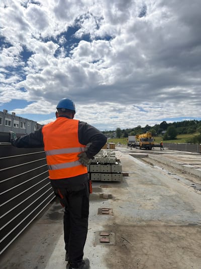 The image depicts a construction site under a cloudy sky, likely in Męcina, Poland. In the foreground, a construction worker wearing a blue hard hat and an orange reflective vest stands with their back to the camera, facing a stack of dark, vertically-oriented panels. The worker's right hand is on their hip, and their left hand rests on the panels. They are also wearing dark work pants and work boots. To the right of the worker, in the midground, are stacks of grey construction materials, possibly pre-cast concrete elements. Further in the background, a yellow crane truck is visible, along with other construction equipment and structures. The ground is concrete, showing some wet patches and debris. The overall scene suggests ongoing construction work.