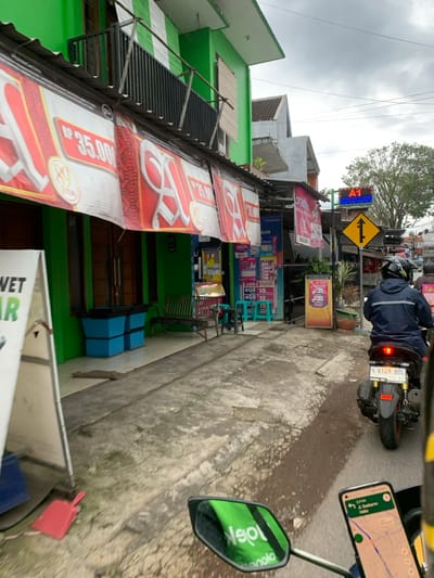 The image shows a street scene in Malang, Indonesia, taken from the perspective of a motorcyclist. The location is at coordinates -7.928 latitude and 112.607 longitude. The street is narrow and paved with concrete and is flanked by buildings on either side. On the left side of the image, there is a green building with a black metal railing in front, and signs advertising "Rp 35.000" and "Rp 35.000" hang from the front of the building. A blue trash can sits near the sidewalk. A wooden bench with a few red chairs can be seen in front of the shop. On the right side of the image, there is a building with various signs and banners. There are many shops and restaurants in the area, and the street is crowded with traffic, including the motorcyclist taking the photo. There is a street sign with an arrow pointing up. The rider is wearing a blue jacket and a helmet. Their motorcycle is parked on the side of the road, and the rear license plate reads "N 6149 ADL". The motorcycle's right side mirror reflects a green sign, possibly a traffic sign, with the word "jol" visible. The photo is taken in daylight, and the sky is cloudy. A smartphone mounted on the motorcycle is visible in the foreground, showing a navigation app with a route and the location "Jl. Sultan Agung" displayed.