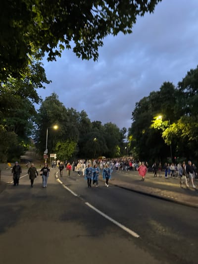 A twilight scene depicts a road in the city of Glasgow, United Kingdom, lined with mature trees on either side. A crowd of people, appearing to be participating in a procession or event, walks down the road. Many individuals are wearing raincoats or ponchos, suggesting inclement weather or a specific event theme. A prominent single street lamp illuminates the scene with a warm yellow light, casting shadows and highlighting the trees and some of the participants. The sky is overcast with dark clouds, contributing to the dim lighting conditions. The road has white lines indicating lanes. Some individuals in the foreground are more clearly visible, while the crowd recedes into the distance. The overall impression is of an organized gathering on a public street under dim, cloudy skies.